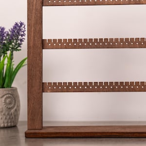 Puede incluir: Un expositor de joyas de madera de color marr&oacute;n oscuro con varias barras horizontales que presentan peque&ntilde;os orificios circulares espaciados uniformemente para colgar pendientes. La estructura tiene una base rectangular plana.