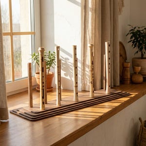 May include: A wooden display stand holding five vertical bamboo flutes of varying lengths. The stand features a tiered, rectangular base with upright supports. Each flute is light tan with dark brown nodes and finger holes arranged along the body.