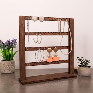 Puede incluir: Un expositor de joyas de madera de color marr&oacute;n oscuro con cuatro barras horizontales que tienen muescas espaciadas uniformemente para colgar pendientes y collares. El soporte tiene una base rectangular y sostiene varios estilos de pendientes y un collar.