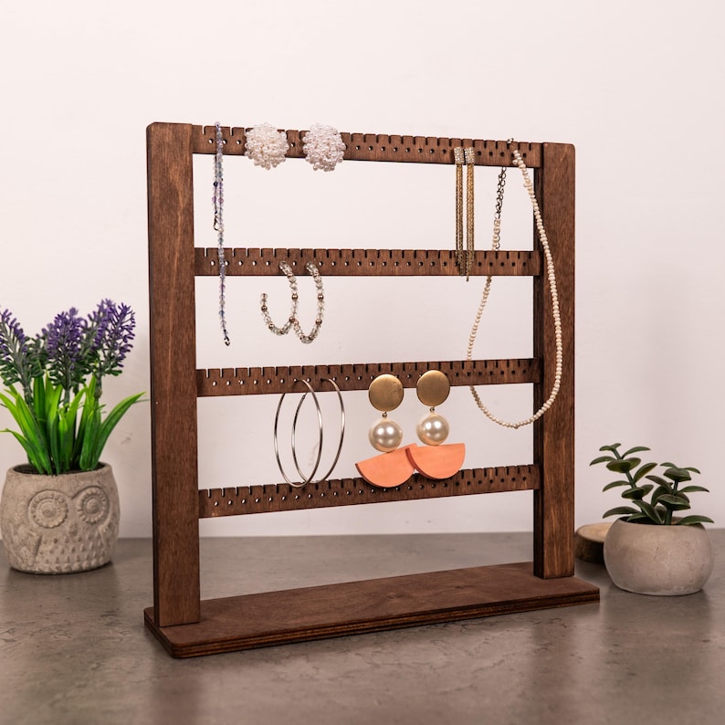 Puede incluir: Un expositor de joyas de madera de color marr&oacute;n oscuro con cuatro barras horizontales que tienen muescas espaciadas uniformemente para colgar pendientes y collares. El soporte tiene una base rectangular y sostiene varios estilos de pendientes y un collar.