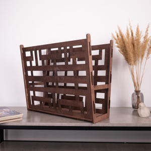May include: A dark brown wooden magazine rack with a geometric design. The rack has a rectangular shape with open sides and a series of horizontal and vertical slats. It is sitting on a gray surface.