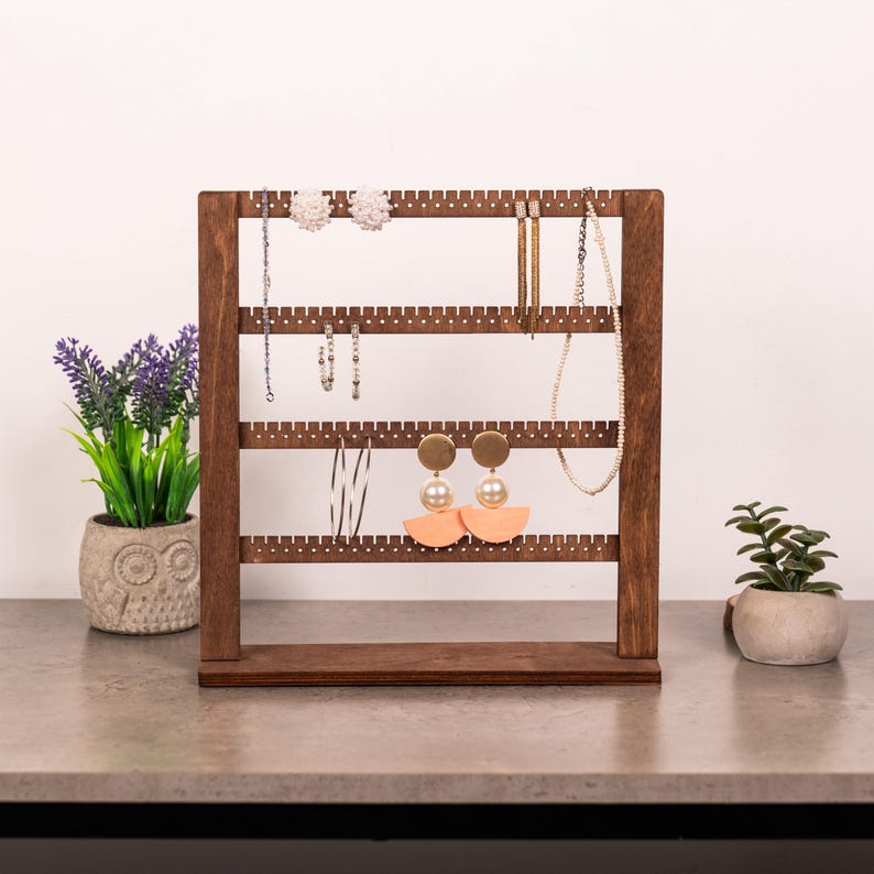 Puede incluir: Un soporte organizador de joyas de madera marr&oacute;n oscuro con cuatro barras horizontales que presentan peque&ntilde;as muescas para colgar objetos. El soporte muestra varios pendientes, incluyendo tachuelas blancas, aros plateados y colgantes.