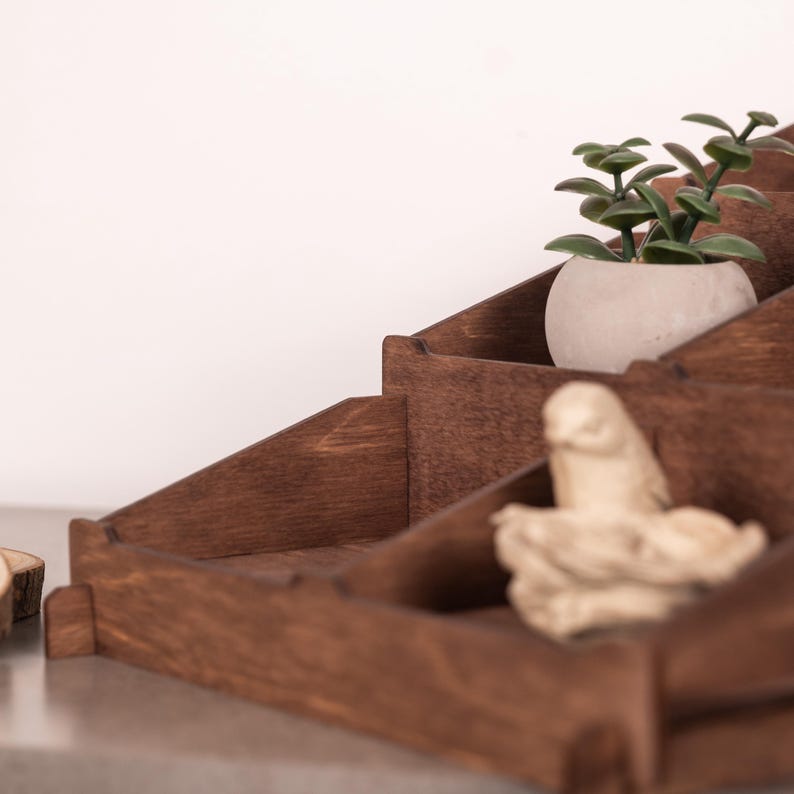 Op de afbeelding: Een getrapte donkerbruine houten displaystandaard met meerdere vakken. Een kleine groene vetplant in een lichtgrijze pot staat in het bovenste gedeelte en een klein lichtgekleurd houten vogelbeeldje rust in het onderste gedeelte.