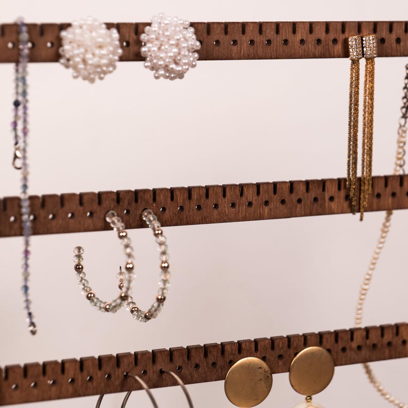 Puede incluir: Un soporte organizador de joyas de madera marr&oacute;n oscuro con tres barras horizontales que tienen muescas y orificios espaciados uniformemente para colgar accesorios. El soporte exhibe varios pendientes, incluyendo racimos tipo perla, flecos dorados, aros con cuentas y discos dorados.