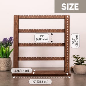 Puede incluir: Un expositor de pendientes de madera marr&oacute;n oscuro con cuatro barras horizontales con peque&ntilde;os orificios para joyer&iacute;a. El soporte mide 25,4 cm de ancho por 26,7 cm de alto, con una base de 7 cm y 4,85 cm de espacio entre las barras.