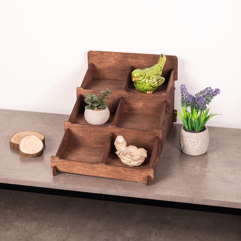 Op de afbeelding: Een getrapte houten displaystandaard met vijf open vakken. De standaard bevat een kleine groene keramische vogel, een witte pot met een groene vetplant en een wit vogelnestbeeldje. Twee ronde houten schijven en een lavendelplant staan ernaast.