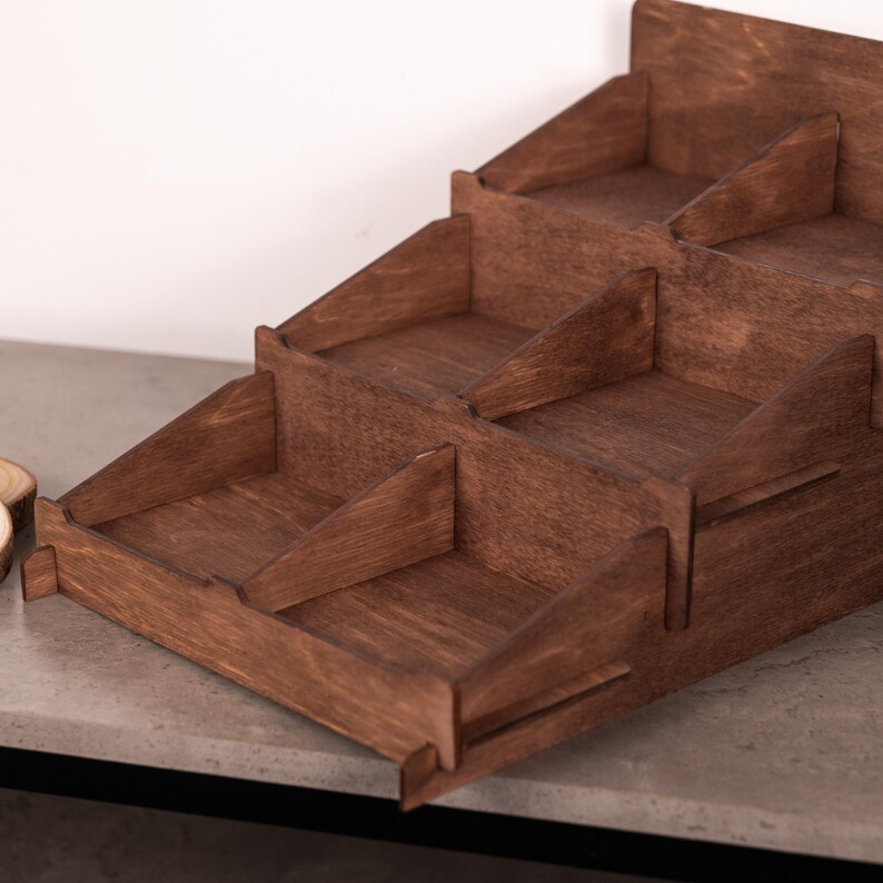 Op de afbeelding: Een getrapte houten displaystandaard met een donkerbruine afwerking en een natuurlijke houtnerftextuur. De structuur bestaat uit drie niveaus, elk verdeeld in meerdere open vakken door middel van verticale houten tussenschotten.