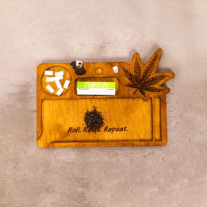 May include: Wooden rolling tray with a carved cannabis leaf design. The tray has compartments for rolling papers, a lighter, and other accessories. The text "Roll. Relax. Repeat." is engraved on the tray.