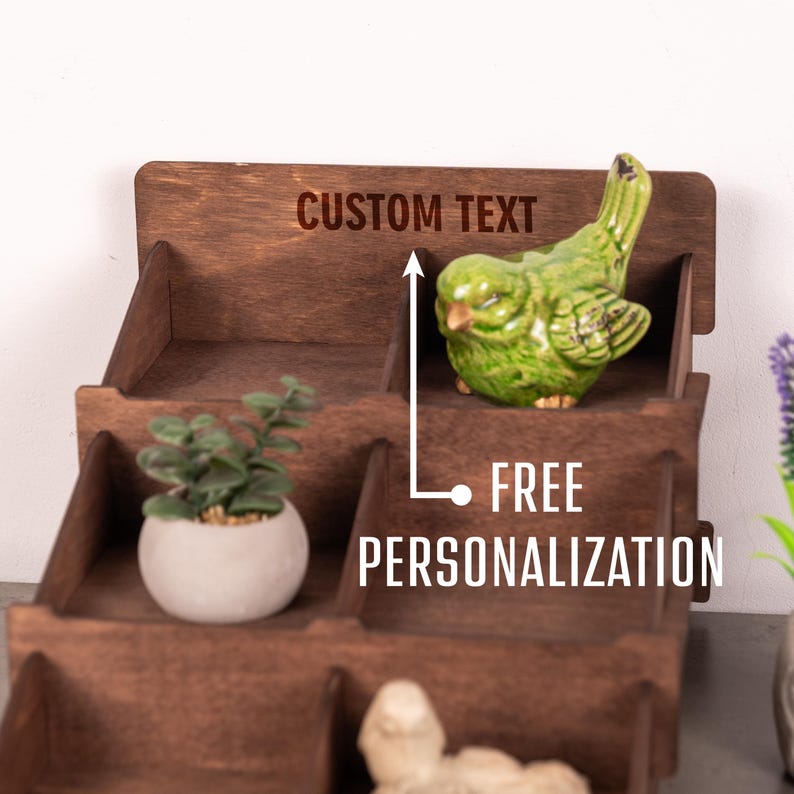 Op de afbeelding: Een getrapte donkerbruine houten displaystandaard met meerdere open vakken. Het achterpaneel bevat de gegraveerde tekst CUSTOM TEXT en een pijl die naar de tekst wijst met het label FREE PERSONALIZATION. Een groen vogelbeeldje en een plantje staan erin.