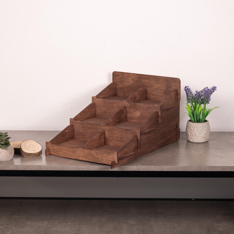 Op de afbeelding: Een donkerbruine houten displaystandaard met een getrapt ontwerp met drie niveaus. Elk niveau is verdeeld in twee aparte compartimenten door verticale houten verdelers, wat in totaal zes open opbergvakken cre&euml;ert voor kleine voorwerpen.