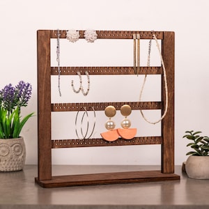 Puede incluir: Un soporte organizador de joyas de madera marr&oacute;n oscuro con cuatro barras horizontales con orificios espaciados uniformemente para colgar pendientes y collares. El marco es rectangular con una base plana y muestra varias piezas de joyer&iacute;a, incluidos pendientes de perlas, aros y un collar de cuentas.