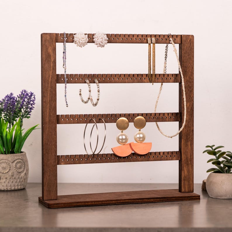 Puede incluir: Un soporte organizador de joyas de madera marr&oacute;n oscuro con cuatro barras horizontales con orificios espaciados uniformemente para colgar pendientes y collares. El marco es rectangular con una base plana y muestra varias piezas de joyer&iacute;a, incluidos pendientes de perlas, aros y un collar de cuentas.