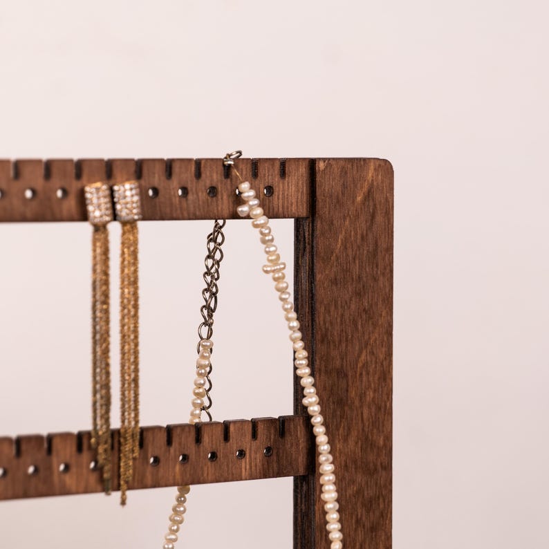 Puede incluir: Un expositor de joyas de madera marr&oacute;n oscuro con listones horizontales que presentan peque&ntilde;as muescas. Colgados del list&oacute;n superior hay dos pendientes colgantes dorados con flecos de cadena y un collar hecho de peque&ntilde;as cuentas blancas y una cadena plateada.