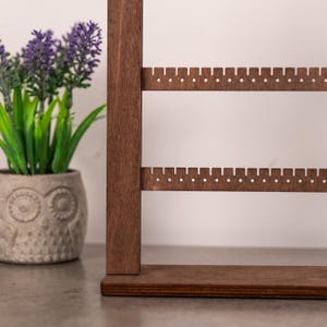 Puede incluir: Un expositor de joyas de madera de color marr&oacute;n oscuro con dos barras horizontales que tienen peque&ntilde;os orificios espaciados uniformemente para colgar pendientes. El soporte cuenta con una base rectangular plana y un poste vertical.