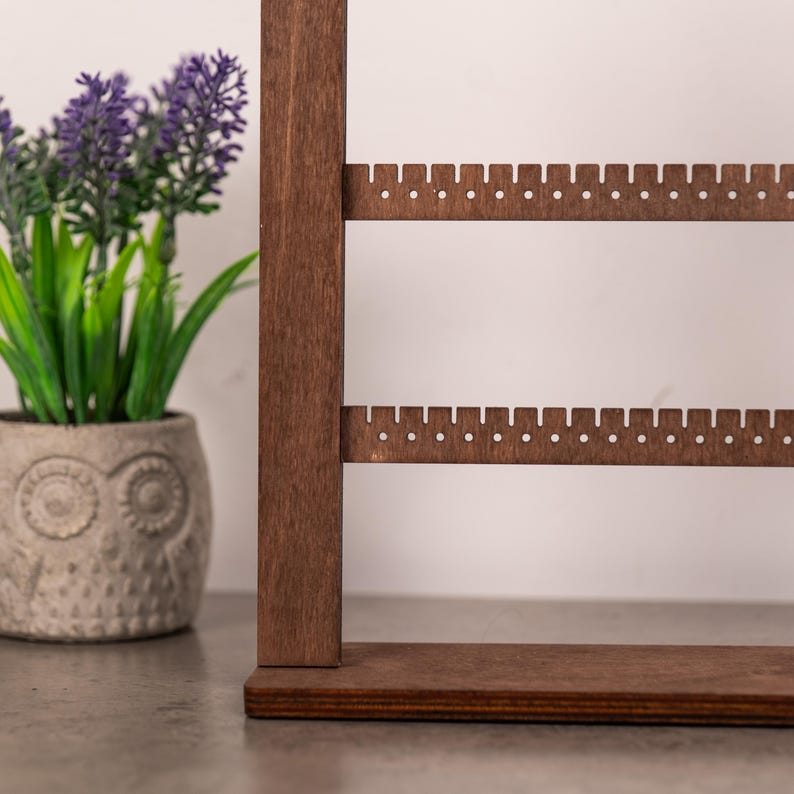 Puede incluir: Un expositor de joyas de madera de color marr&oacute;n oscuro con dos barras horizontales que tienen peque&ntilde;os orificios espaciados uniformemente para colgar pendientes. El soporte cuenta con una base rectangular plana y un poste vertical.
