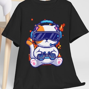 May include: Black t-shirt featuring a pixelated cartoon cat wearing a VR headset and holding a game controller. The cat is white with orange accents. The background has orange and red pixelated elements. A white baseball cap hangs on a wooden chair.