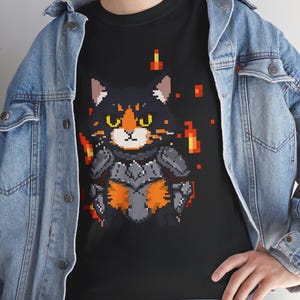 Pode incluir: Camiseta preta com um design de gato guerreiro pixelizado. O gato tem pelo preto, olhos laranja e usa uma armadura cinza. Chamas e detalhes pixelizados cercam o gato, criando uma camiseta gráfica única.