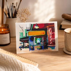 Puede incluir: Una impresión artística rectangular que presenta una ilustración estilizada y colorida del exterior de un escaparate con un toldo rojo, un letrero amarillo y un poste de barbero. La obra utiliza bloques de colores vivos como verde, azul y rojo.