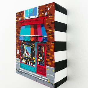 Puede incluir: Un cuadro rectangular sobre lienzo que representa un escaparate con fachada de ladrillo, un toldo de rayas multicolores y una puerta roja. Los bordes laterales del lienzo están pintados con gruesas rayas horizontales blancas y negras.