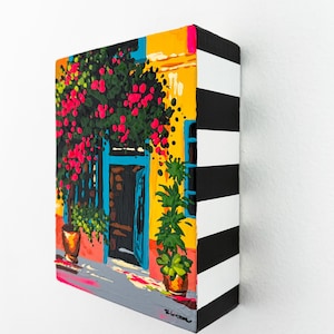 Puede incluir: Un cuadro rectangular sobre lienzo que muestra la fachada de un edificio amarillo con una puerta azul brillante, flores rosas vibrantes y plantas verdes en macetas. Los bordes laterales del lienzo están pintados con rayas negras y blancas.