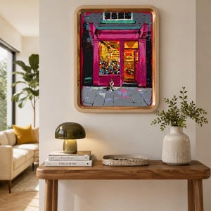 Puede incluir: Un cuadro rectangular enmarcado que representa un escaparate con una fachada de color magenta vibrante y un gran ventanal. Un perro pequeño está sentado en la acera gris frente a la tienda. La pintura presenta pinceladas audaces y brillantes.