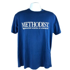 May include: A navy blue t-shirt featuring the white text "METHODIST" across the chest, with "METHODIST HOSPITAL OF JONESBORO" printed below. The t-shirt has short sleeves and is a classic crew neck style.