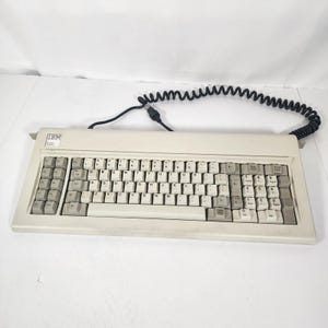 May include: A beige IBM computer keyboard with a QWERTY layout, featuring light and dark grey keys, a numeric keypad, and a black coiled cable. The IBM logo is printed in the upper left corner of the rectangular plastic casing.