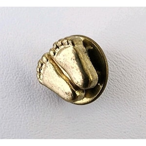 Puede incluir: Un broche dorado con un par de huellas de bebé. Las huellas están detalladas y parecen estar ligeramente elevadas de la superficie del broche. El broche tiene una base circular.