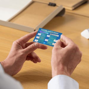 May include: A blue and white Suture Quick Guide card is held between two hands. The card has text and diagrams. The background is a light brown desk with a white object and a pen holder.