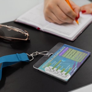 May include: A clear plastic badge holder with a blue lanyard and silver clasp. The badge has text that reads "NEEDLES & OR CALL-OUTS" and includes a list of needle types. A pair of glasses and a notebook are also visible.