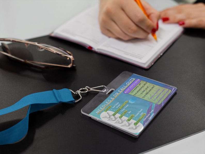 May include: A clear plastic badge holder with a blue lanyard and silver clasp. The badge has text that reads "NEEDLES & OR CALL-OUTS" and includes a list of needle types. A pair of glasses and a notebook are also visible.