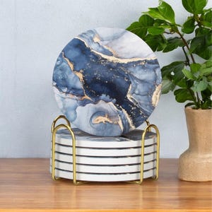 Puede incluir: Juego de seis posavasos redondos con diseño de mármol azul y dorado, exhibidos en un soporte de metal dorado. Los posavasos tienen un acabado brillante y están apilados sobre una superficie de madera. Una planta en una maceta beige está en el fondo.