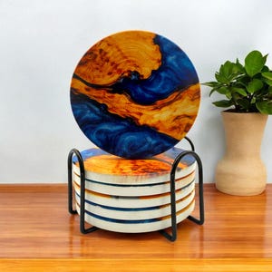 May include: A set of six round coasters with a unique design. The coasters feature a blend of blue and gold resin, resembling wood grain. They are displayed in a black metal holder, perfect for protecting surfaces from drink rings. The coasters are approximately 4 inches in diameter.