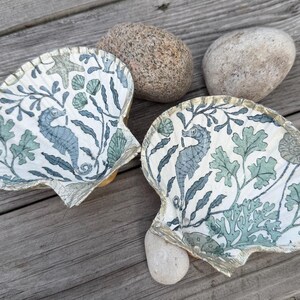 Puede incluir: Dos conchas decorativas con un diseño de caballito de mar y botánico en azul y verde sobre fondo blanco. Los bordes son dorados. Dos piedras están colocadas cerca.