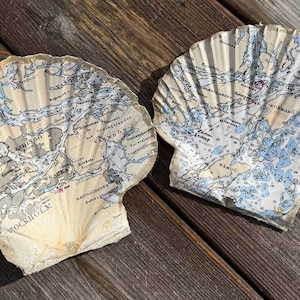 May include: Two large, scallop-shaped decorative pieces with a vintage map design. The maps feature blue, grey, and beige tones, with text indicating locations. The pieces are placed on a weathered wooden surface.