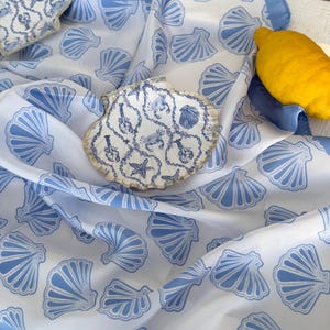 Könnte beinhalten: Ein weißes und blaues Textil mit Muschelmotiv und blauem Rand. Der Stoff ist mit blauen Muscheln gemustert. Zwei muschelförmige Dekorationsgegenstände sind sichtbar, zusammen mit einer gelben Zitrone. Das Gesamtthema ist küsten- und nautisch.