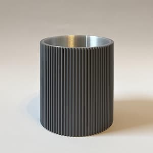 Könnte beinhalten: Ein zylindrischer Behälter mit geriffelter, dunkelgrauer Außenseite und silberfarbenem Innenraum. Die vertikalen Rillen erzeugen eine strukturierte Oberfläche. Der Behälter steht vor einem schlichten weißen Hintergrund.