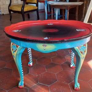 Peut inclure: Une table d'appoint peinte à la main avec un plateau rouge et noir et une base turquoise. La table présente des pieds incurvés ornés avec des accents à carreaux et des détails floraux décoratifs. La table mesure environ 61 cm de diamètre et 51 cm de haut.