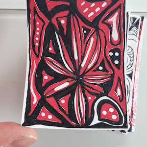 Può includere: Una carta rettangolare con un'illustrazione disegnata a mano che presenta un motivo floreale centrale a forma di stella in rosso, nero e bianco. Il design include contorni neri spessi, forme geometriche rosse e nere e piccoli punti bianchi sparsi.
