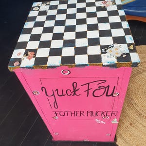 Peut inclure: Une table d'appoint carrée avec un plateau à damier noir et blanc orné de décalcomanies florales aux coins. Les côtés sont peints en rose vif avec le texte manuscrit noir Yuck Fou Fother Mucker et des accents circulaires sur les bords.
