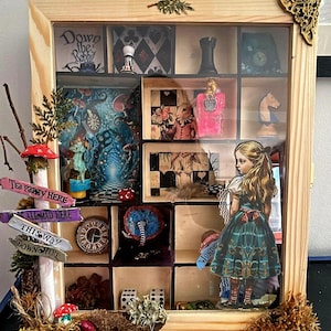 Może przedstawiać: Drewniane pudełko cieni z motywem Alicji w Krainie Czarów. Pudełko zawiera miniaturowe sceny i figurki, w tym Alicję, królika, zegar i znaki z napisem "Tea Party Here" i "This Way Down Here".
