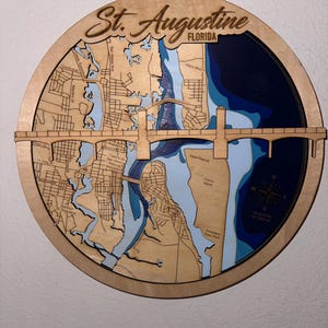 May include: A round wooden map of St. Augustine, Florida, with a bridge and waterways. The map is laser-cut with intricate details, including street names and depth contours. The text "St. Augustine FLORIDA" is at the top.