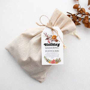 Op de afbeelding: Een beige stoffen zakje, vastgebonden met touw, met een vakantie potpourri. Een wit label is bevestigd, met de tekst "Holiday Simmering Potpourri" en instructies. Het label is versierd met feestelijke botanische illustraties.