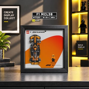 May include: A black framed display featuring a LEGO McLaren F1 MCL38 race car model and a matching driver minifigure. The background has a white and orange graphic with the LEGO and McLaren logos, plus the text MCL38 and Formula One branding.