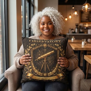 Peut inclure: Un coussin décoratif avec un fond marron foncé représentant une illustration de télescope doré. Le texte "THE ROYAL OBSERVATORY" est en haut, avec "EXPLORARE ASTRA" en bas. Le coussin est tenu par une personne.