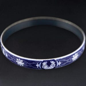 May include: A blue and white enamel bangle bracelet. The bracelet features a dark blue background with a repeating floral pattern in white. The metal band has a silver finish. The bracelet is a circular shape.