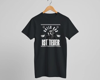 Könnte beinhalten: Schwarzes T-Shirt mit einer weißen Grafik eines Radfahrers und dem deutschen Text "Guter Rad ist Teuer". Das Design beinhaltet ein Schachbrettmuster und stilisierten Text. Das Shirt hängt an einem Holzbügel.