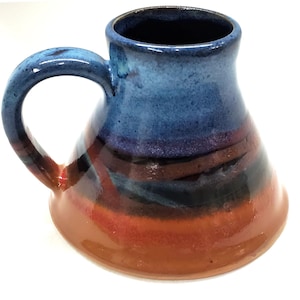 May include: A handmade ceramic mug with a unique, flared shape. The mug features a gradient of colors, including blue, purple, black, and red-orange. It has a large, curved handle and a glossy finish, perfect for coffee or tea.