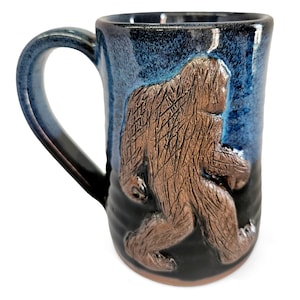 May include: A ceramic mug with a blue and black glaze, featuring a brown Bigfoot figure in relief. The mug has a large handle and a capacity suitable for coffee or tea.