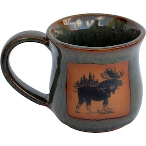 May include: A ceramic coffee mug with a dark green glaze and a brown handle. The mug features a rectangular design with a black illustration of a moose and trees. The mug is designed for hot beverages.